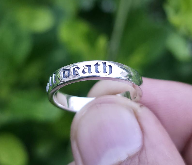 May include: A silver ring with the word "death" engraved in a gothic font. The ring is held in a hand against a blurred green background. The ring has a polished finish.