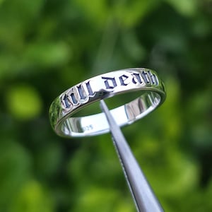 May include: A silver ring with the words "till death" engraved in a gothic font. The ring is held by a pair of tweezers against a blurred green background. The ring is a band style, with a polished finish.