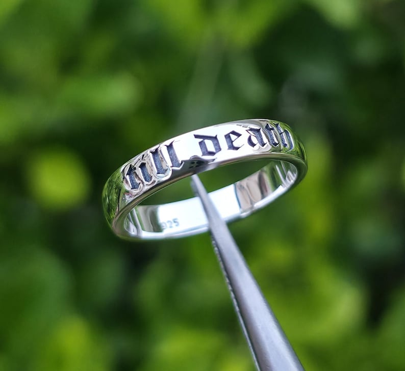 May include: A silver ring with the words "till death" engraved in a gothic font. The ring is held by a pair of tweezers against a blurred green background. The ring is a band style, with a polished finish.