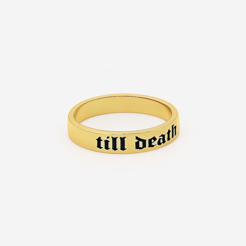 May include: A gold-colored ring with the phrase "till death" in black gothic lettering. The ring is a simple band design, with a smooth, polished surface. The ring is set against a plain white background.