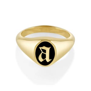 Puede incluir: Un anillo de sello dorado con un centro ovalado negro con la letra "A" en fuente gótica. El anillo tiene un acabado pulido y está sobre un fondo blanco. Este anillo es una joya.