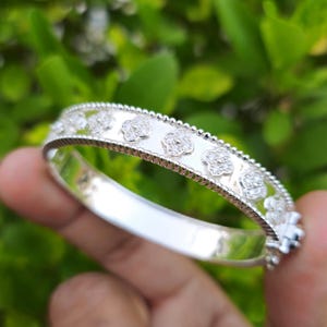 May include: A silver bangle bracelet with a floral design. The bracelet features a series of raised, flower-shaped embellishments set with small, sparkling stones. The edges of the bracelet are detailed with a delicate, beaded pattern. The bracelet is shown against a blurred green background.