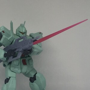 May include: A green and gray robot action figure holding a gun with a pink beam of light coming out of the end of the barrel.