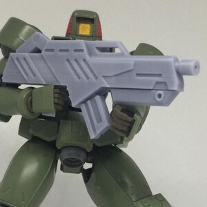 May include: A green action figure holding a gray rifle. The figure has a red helmet and is posed as if ready for battle. The rifle has a detailed design with a barrel and a handle. The figure is in a combat stance.