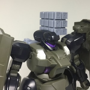 May include: A black and green robot toy with a gray block accessory on its head. The robot has a purple visor and white accents.
