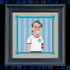 May include: Cross-stitch portrait of a person with brown hair, wearing glasses, a white shirt with a red trim and a blue 'M' on the chest, and a pink and black bracelet.
