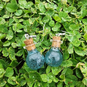 May include: A pair of blue glass bottle earrings with cork stoppers and silver hooks. The earrings are on a green background.
