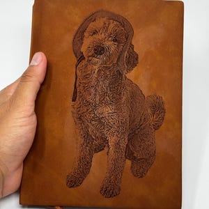 May include: A brown leather journal featuring an engraved poodle wearing a hat. The journal has a matching brown ribbon bookmark. The cover is held by a hand.