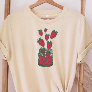 May include: A cream colored t-shirt with a graphic of a glass jar filled with red strawberries and red strawberries flying out of the jar.