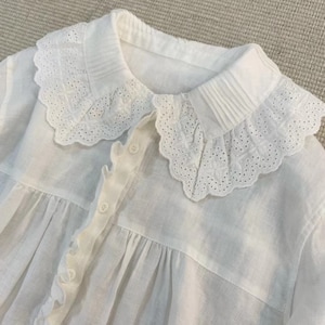 Peut inclure: Chemise blanche en coton avec un grand col en dentelle et une fermeture boutonnée à l'avant. La chemise a un corsage plissé et des manches longues.
