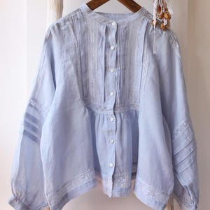 May include: A light blue long-sleeve blouse featuring a mandarin collar, a front button placket, and vertical pintuck pleats. The garment is accented with white lace trim along the chest, shoulders, and hem, with additional pleating on the sleeves.