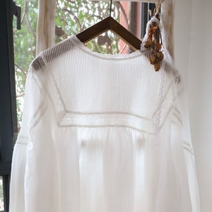 May include: White cotton blouse with a lace trim and a gathered neckline. The blouse has long sleeves with a gathered cuff.