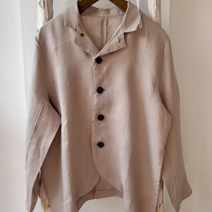 Può includere: Un blazer in lino beige con colletto a revers e quattro bottoni neri. La giacca a maniche lunghe è appesa a una gruccia di legno su sfondo bianco. Il blazer ha una vestibilità rilassata e un orlo curvo unico.
