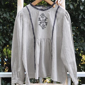May include: A long-sleeved blouse featuring a fine grey and white pinstripe pattern, a round neckline with dark grey lace trim, and a central floral embroidery design. The garment includes vertical dark grey lace accents and gathered fabric details.