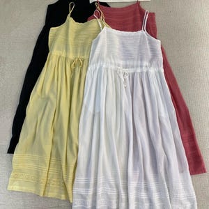 Puede incluir: Cuatro vestidos de tirantes finos en negro, amarillo, blanco y rojo. Los vestidos tienen cintura con cordón y un ribete de encaje en el dobladillo. Los vestidos están colgados en una percha blanca.