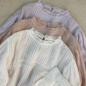 Puede incluir: Tres blusas de manga larga en colores pastel: blanco, melocotón y lavanda. Cada blusa presenta un cuello alto con delicados adornos de encaje y pliegues verticales. Las mangas son largas y ligeramente abullonadas.