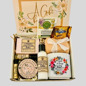 Puede incluir: Una caja de regalo llena de artículos de cuidado personal. Incluye jabón de leche de cabra, loción corporal, una vela, una taza floral, bálsamo labial, una bolsita de té, una barra de chocolate y un paño. La caja está decorada con motivos florales.