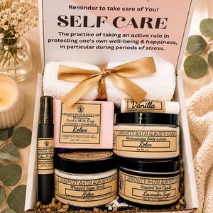 May include: A self-care gift box containing a white towel with a gold ribbon, a 4 oz bar of lavender and chamomile goat's milk soap, a 5 oz jar of body lotion, a jar of whipped sugar scrub, a 4 oz lavender sage soy candle, and a vanilla lip balm.