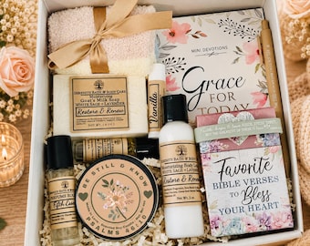 May include: A gift box containing a fuzzy beige cloth with a gold ribbon, a bar of goat's milk soap, a vanilla lip balm, a perfume roller, a scented candle labeled Be Still & Know, a body lotion, a devotional book, a pen, and a verse card set.