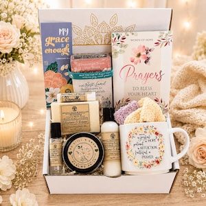 May include: A gift box containing a vanilla lip balm, a goat's milk soap bar, a body lotion, a roll-on perfume, a candle tin labeled Be Still and Know, a mug with floral text, a prayer book, and a bookmark with the text My Grace Is Enough.