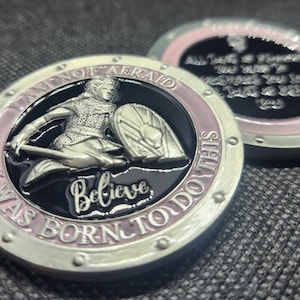 May include: Two silver-colored round coins with a pink and black design. One coin features a woman in armor holding a sword and shield with the text "I am not afraid I was born to do this Believe". The other coin features the text "All things are possible to him who believes in me".