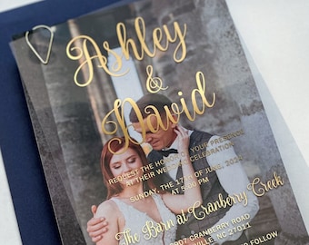 May include: A clear acrylic wedding invitation with gold foil lettering. The invitation features a photo of a couple and the text "Ashley & David" and "The Barn at Cranberry Creek".