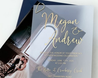 May include: Wedding invitation with a translucent, light blue background and gold lettering. The invitation reads "Megan & Andrew" and includes the date, time, and location of the wedding ceremony. The Barn at Cranberry Creek, 2907 Cranberry Road, Boonville, NC 27011. Dinner and dancing to follow.