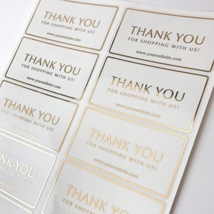 Puede incluir: Una hoja de pegatinas blancas con letras doradas. Las pegatinas dicen "¡Gracias por comprar con nosotros!" e incluyen una dirección de sitio web.