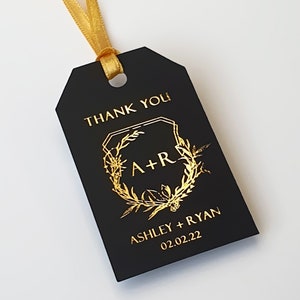 May include: A rectangular black gift tag with a gold ribbon loop. The tag features gold foil text that reads THANK YOU, A+R, ASHLEY + RYAN, and 02.02.22, centered within a gold foil botanical wreath design with geometric border elements.