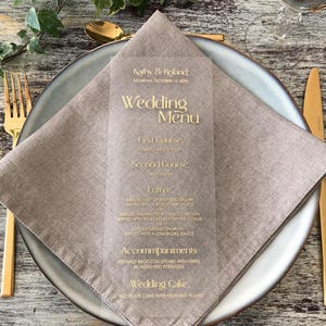 Op de afbeelding: Een helder acryl trouwmenu met gouden letters, met gerechten zoals tomatensoep en gegrilde zalm. Het menu ligt op een gevouwen beige linnen servet, op een grijze plaat met gouden bestek. Het menu is gepersonaliseerd voor Kathy & Roland.