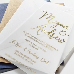 May include: A white wedding invitation with gold lettering. The invitation reads "Megan & Andrew" and includes the date, time, and location of the wedding. The text reads "Request the honour of your presence at their wedding celebration. Sunday, June 17th, 2024 at 5:00 PM. The Barn at Cranberry Creek, 2907 Cranberry Road, Boonville, NC 27011. Dinner and dancing to follow."