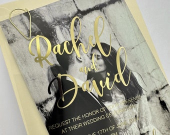 May include: Wedding invitation featuring a black and white photo of a couple kissing, with gold foil lettering that reads "Rachel and David." The invitation includes the wedding date, time, and location. A gold paperclip holds the invitation.