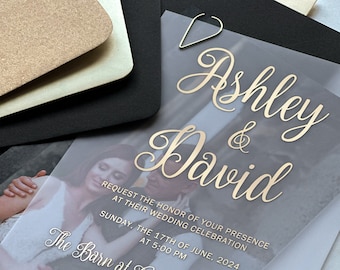 May include: A clear plastic wedding invitation with gold lettering that reads "Ashley & David". The invitation includes a photo of the couple and details about the wedding ceremony, which will be held at The Barn at Cranber on Sunday, June 17th, 2024 at 5:00 PM.
