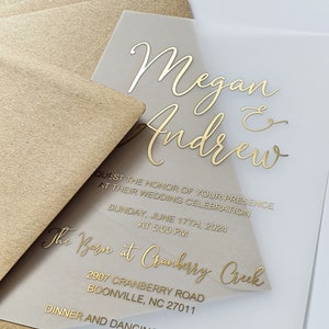 May include: A clear acrylic wedding invitation with gold foil lettering. The invitation reads "Megan & Andrew" and includes the details of their wedding celebration at "The Barn at Cranberry Creek" on Sunday, 17th June 2024 at 5:00 PM. The address is 2907 Cranberry Road, Boonville, NC 27011.