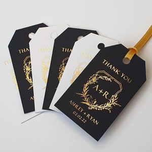 May include: A set of rectangular black and white gift tags featuring gold foil lettering that reads THANK YOU, A+R, ASHLEY + RYAN, and 02.02.22. Each tag has a circular hole at the top, and one black tag is threaded with a gold ribbon.