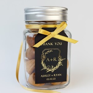May include: A clear glass jar with a silver metal lid filled with brown treats. A black rectangular tag is tied to the jar with a gold ribbon. The tag features gold text reading THANK YOU, A+R, ASHLEY + RYAN, and the date 02.02.22.