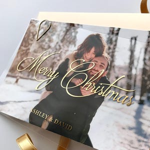 Op de afbeelding: Een kerstkaart met een foto van een stel in een besneeuwde buitenomgeving. De woorden "Merry Christmas" staan in gouden letters, met de namen "Ashley & David" eronder. Een gouden lint en een gouden paperclip zijn ook zichtbaar.