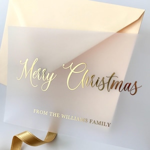 May include: A translucent Christmas card with gold foil lettering that reads "Merry Christmas" and "FROM THE WILLIAMS FAMILY." The card is paired with a cream-colored envelope and a gold ribbon.