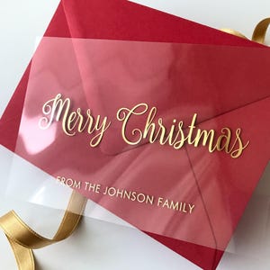 Może przedstawiać: Czerwona koperta z przezroczystą nakładką z napisem "Merry Christmas" i "FROM THE JOHNSON FAMILY" w kolorze złotym. Złota wstążka widoczna w tle. Kartka świąteczna.