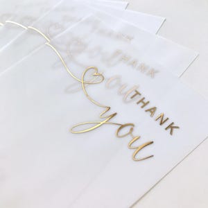 May include: A stack of white translucent cards featuring gold foil lettering that reads "Thank You". The words are in a cursive font with a heart-shaped design. The cards are slightly overlapping, with the top card in focus.