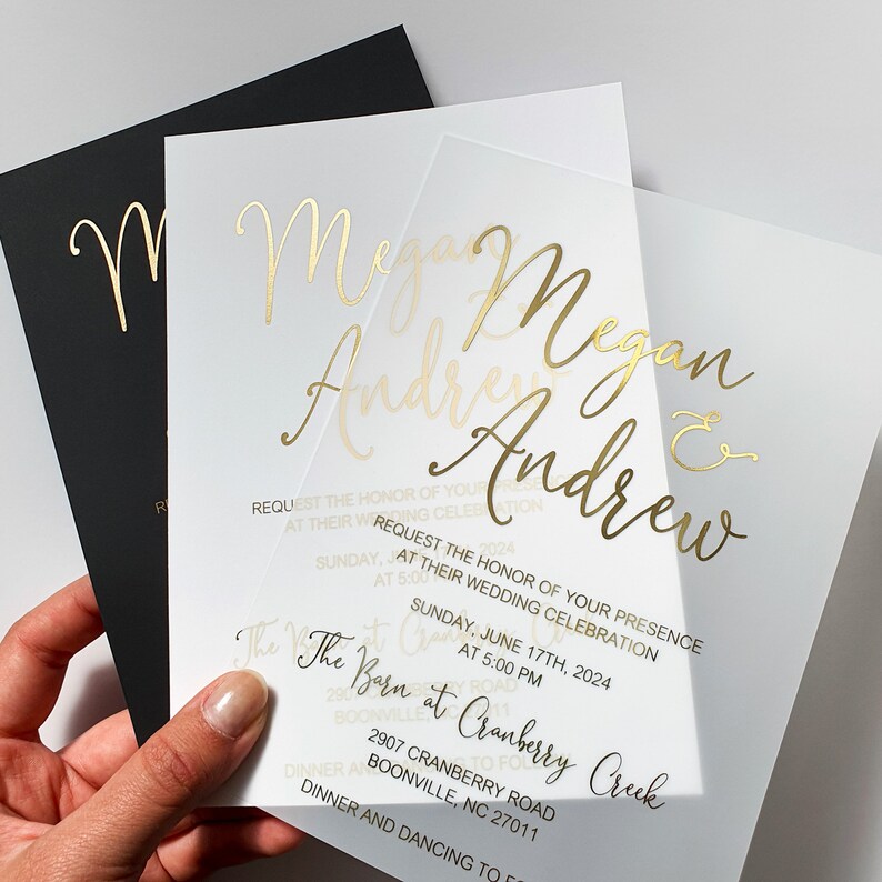 May include: A clear acrylic wedding invitation with gold foil lettering. The invitation reads "Megan & Andrew" and includes the date and time of the wedding, the location, and the text "The Barn at Cranberry Creek".