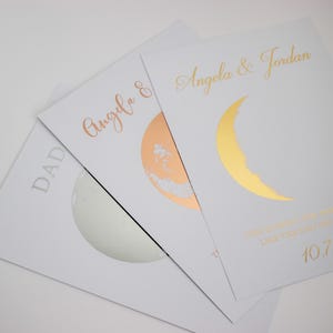 May include: Three white cards with metallic accents. One card has "DAD" in silver. Another has "Angela &" in rose gold with a rose gold moon. The third card has "Angela & Jordan" and a gold crescent moon.
