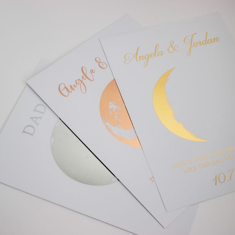 May include: Three white cards with metallic accents. One card has "DAD" in silver. Another has "Angela &" in rose gold with a rose gold moon. The third card has "Angela & Jordan" and a gold crescent moon.