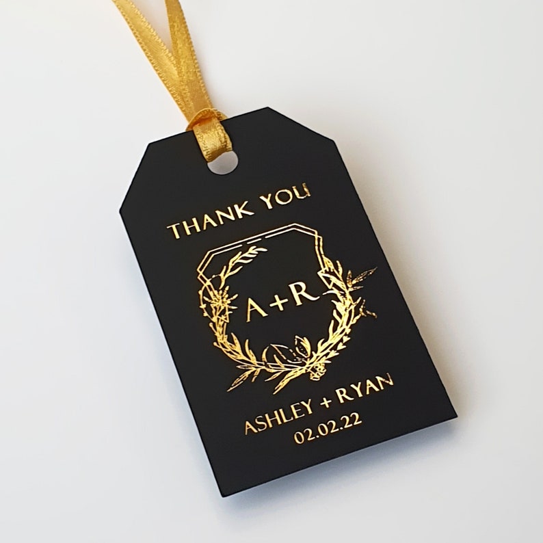 May include: A rectangular black gift tag with a gold ribbon loop. The tag features gold foil text that reads THANK YOU, A+R, ASHLEY + RYAN, and 02.02.22, centered within a gold foil botanical wreath design with geometric border elements.