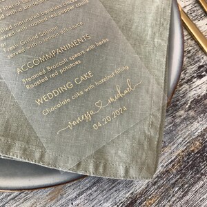 May include: A clear acrylic wedding menu card with gold lettering. The menu includes the names Vanessa and Michael, the date 04.20.2024, and the wedding cake selection: Chocolate cake with hazelnut filling.