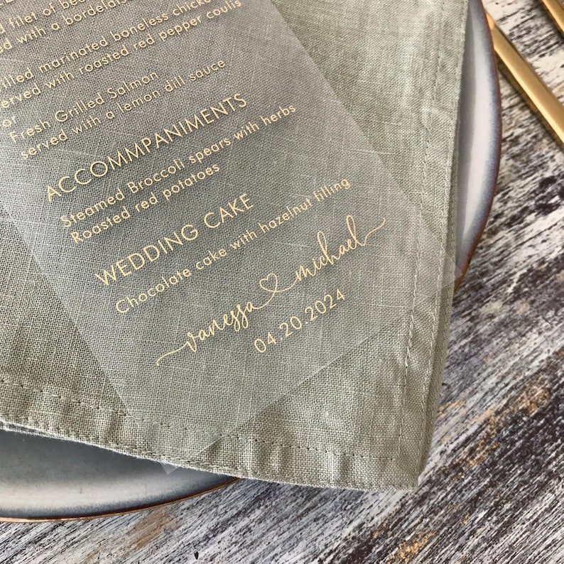 May include: A clear acrylic wedding menu card with gold lettering. The menu includes the names Vanessa and Michael, the date 04.20.2024, and the wedding cake selection: Chocolate cake with hazelnut filling.