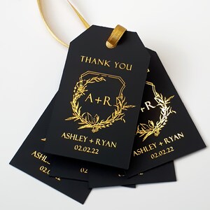 May include: A set of rectangular black gift tags with gold foil lettering and a gold ribbon. The tags feature the text THANK YOU, the initials A+R inside a botanical wreath, the names ASHLEY + RYAN, and the date 02.02.22 printed in gold.