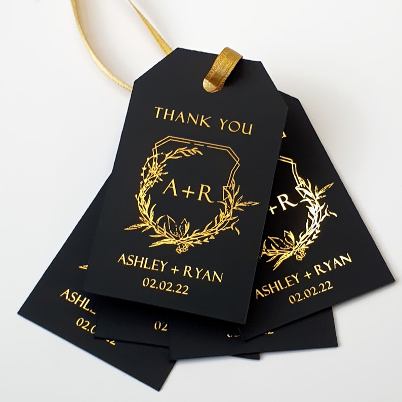 May include: A set of rectangular black gift tags with gold foil lettering and a gold ribbon. The tags feature the text THANK YOU, the initials A+R inside a botanical wreath, the names ASHLEY + RYAN, and the date 02.02.22 printed in gold.
