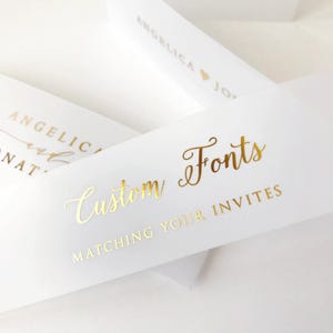 Peut inclure: Cartes d'invitation blanches avec lettrage doré. Le texte sur les cartes indique "Custom Fonts" et "Matching Your Invites". D'autres cartes portent le nom "Angelica".
