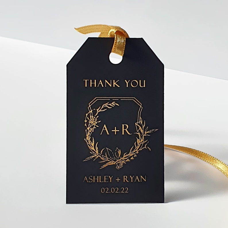 May include: A rectangular black gift tag featuring gold foil text that reads THANK YOU, A+R, ASHLEY + RYAN, and 02.02.22. A gold floral wreath design surrounds the initials. A gold ribbon is threaded through a hole at the top of the tag.